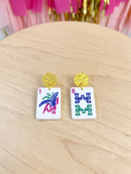 A pair of earrings shaped like white mahjong tiles with gold studs, displayed on a light wood surface with pink tinsel in the background.