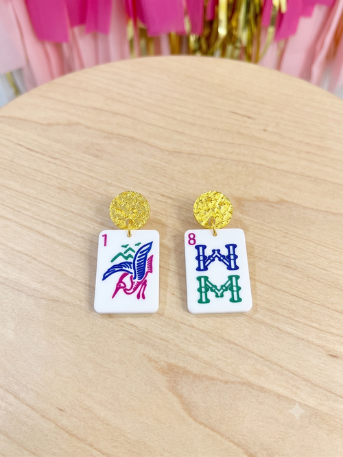 A pair of earrings shaped like white mahjong tiles with gold studs, displayed on a light wood surface with pink tinsel in the background.