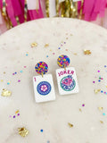 A pair of novelty earrings shaped like playing card tiles, featuring a Joker card on one side and a 1 of Clubs card on the other, with colorful confetti stud tops, displayed on a marble surface with gold confetti.