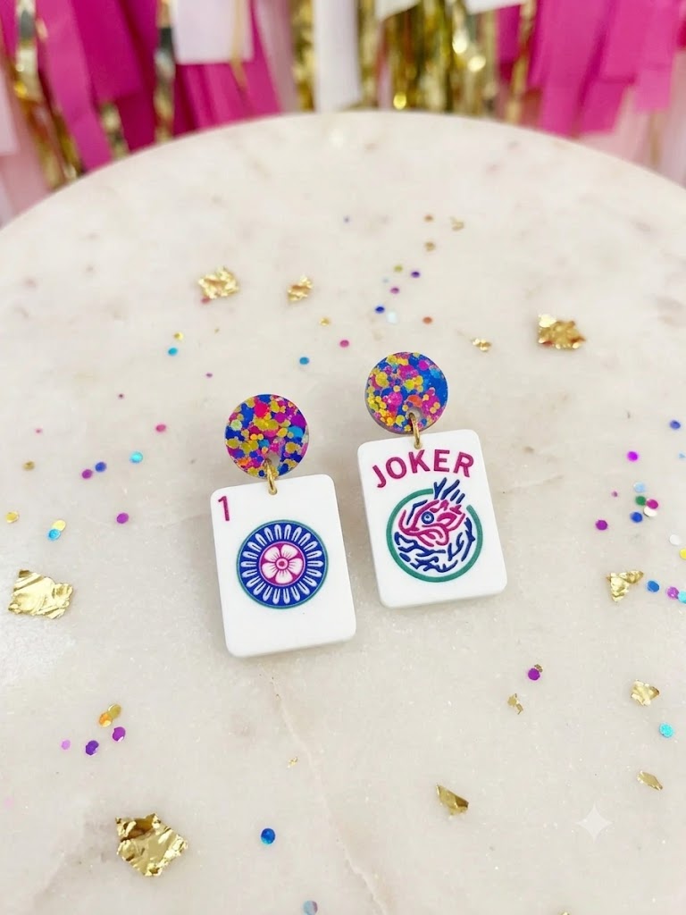 A pair of novelty earrings shaped like playing card tiles, featuring a Joker card on one side and a 1 of Clubs card on the other, with colorful confetti stud tops, displayed on a marble surface with gold confetti.